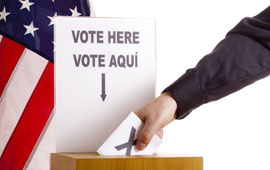 hand inserting ballot into box under bilingual English/Spanish sign to vote with U.S. flag in background