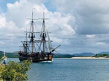 A three-masted sailing ship with its sails furled, sitting motionless on a body of water. A beach and forested hills are in the background.