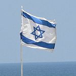 A rectangular flag, white background, blue bands close to the top and bottom, with a blue star of David in the middle. The flag is on a flagpole, with blue sky and sea in the background.