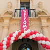 Vertical welcome banner hanging from building balcony above red and white balloon arch