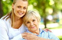 Young female caregiver smiling and embracing an elderly woman sitting on chair in park.