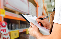 Person holding clipboard and pen, checking warehouse shelf inventory.