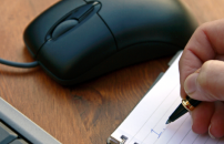 Closeup of person's hand taking notes next to laptop and computer mouse.