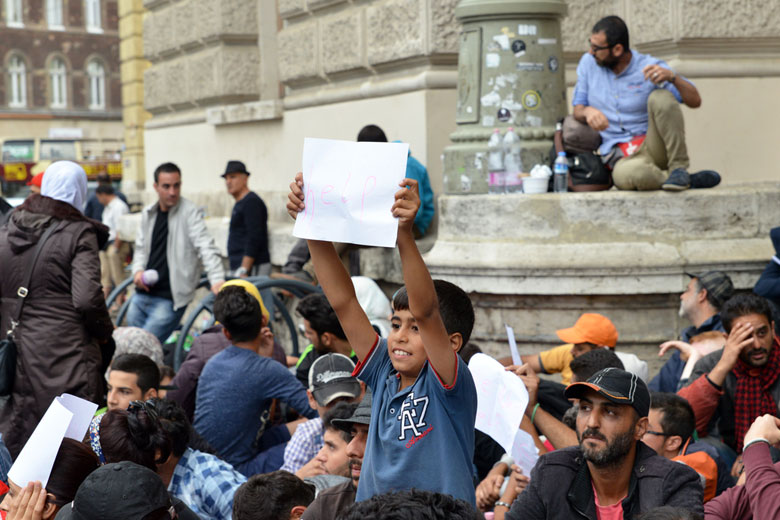Boy holding a 'help' sign surrounded by many other refugees. 