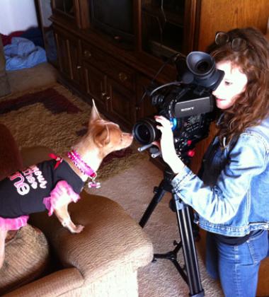 image of filmmaker Melissa Langer holding a film camera which is pointed at a chiuahua wearing a dress.