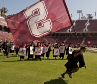 photo of student in cap and gown running across stadium field with Stanford flag