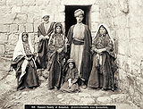 Three women and two men, all sporting serious expressions are standing together with a young girl who is sitting on the floor. The men - one a young man and the other an older man with a white beard - and one of women are looking directly at the camera, while the girl appears to be blinking and laughing. The men are both wearing turbans and striped thobs (dresses) with a robe overtop that is open at the front. The women are wearing dresses that are heavily embroidered over the chest area and overtop of these a type of jacket. All of the women and the girl are wearing veils on their heads that show some of their hair at the front and extend midway down their backs. The girl's headdress includes coins across the top of her head that appear to be attached to the rim of the veil. The caption reads: "618 Peasant Family of Ramallah", followed by the same in German.