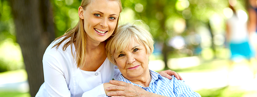 Young female caregiver smiling and embracing an elderly woman sitting on chair in park.