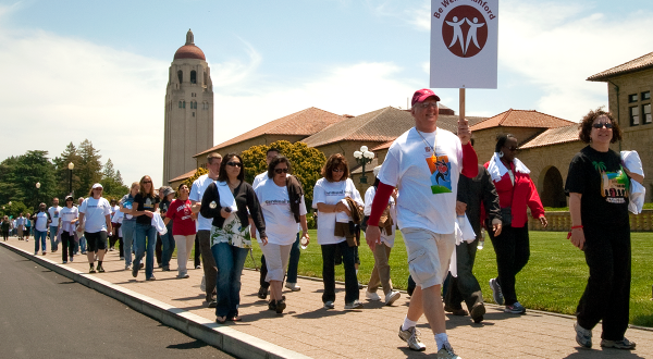 Stream of staff participating in the annual Cardinal Walk on The Oval sidewalk, Hoover Tower in background, with man in front holding a "BeWell@Stanford" sign.