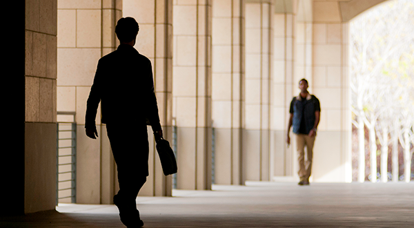 Silhouette of person in foreground carrying bag or briefcase walking away through hallway, with male in background walking forward