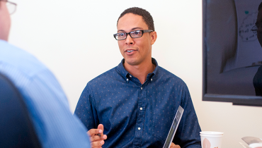Young African American male wearing glasses, pen in hand, talking to male colleague