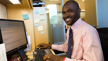 Male African American staffer at desktop computer in office, smiling at viewer