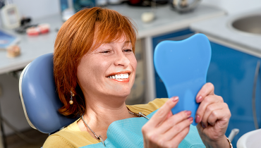 Senior woman with red hair looking at her smile in tooth-shaped mirror at dentist's office