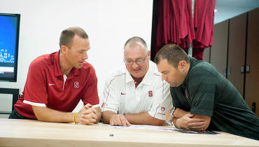 Three male Athletics colleagues huddling over table in discussion.