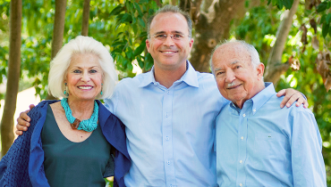 Male staff member with arms around elderly parents with trees in background