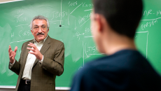 Professor with grayish hair, glasses and moustache, standing in front of green chalkboard, delivering a lecture to students.