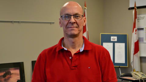 Un homme photographié dans un bureau, deux drapeaux canadiens derrière lui 