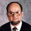 man with dark brown hair, short brown beard, and large glasses. Man is wearing a suit.