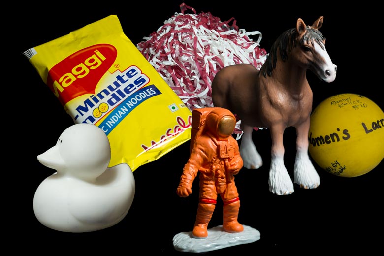 A collage of items including  a Cardinal and white pon pon; a bag of Indian noodles, a white rubber ducky; a women's lacrosse ball; a plasitc horse and a toy astronaut