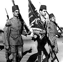 The sides of three men in military uniform moving to the right. The middle one is holding a flag, while the others are holding rifles.