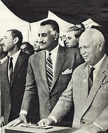 Three men in the foreground looking slightly down. They are all wearing suits and the one on the right is highly decorated.