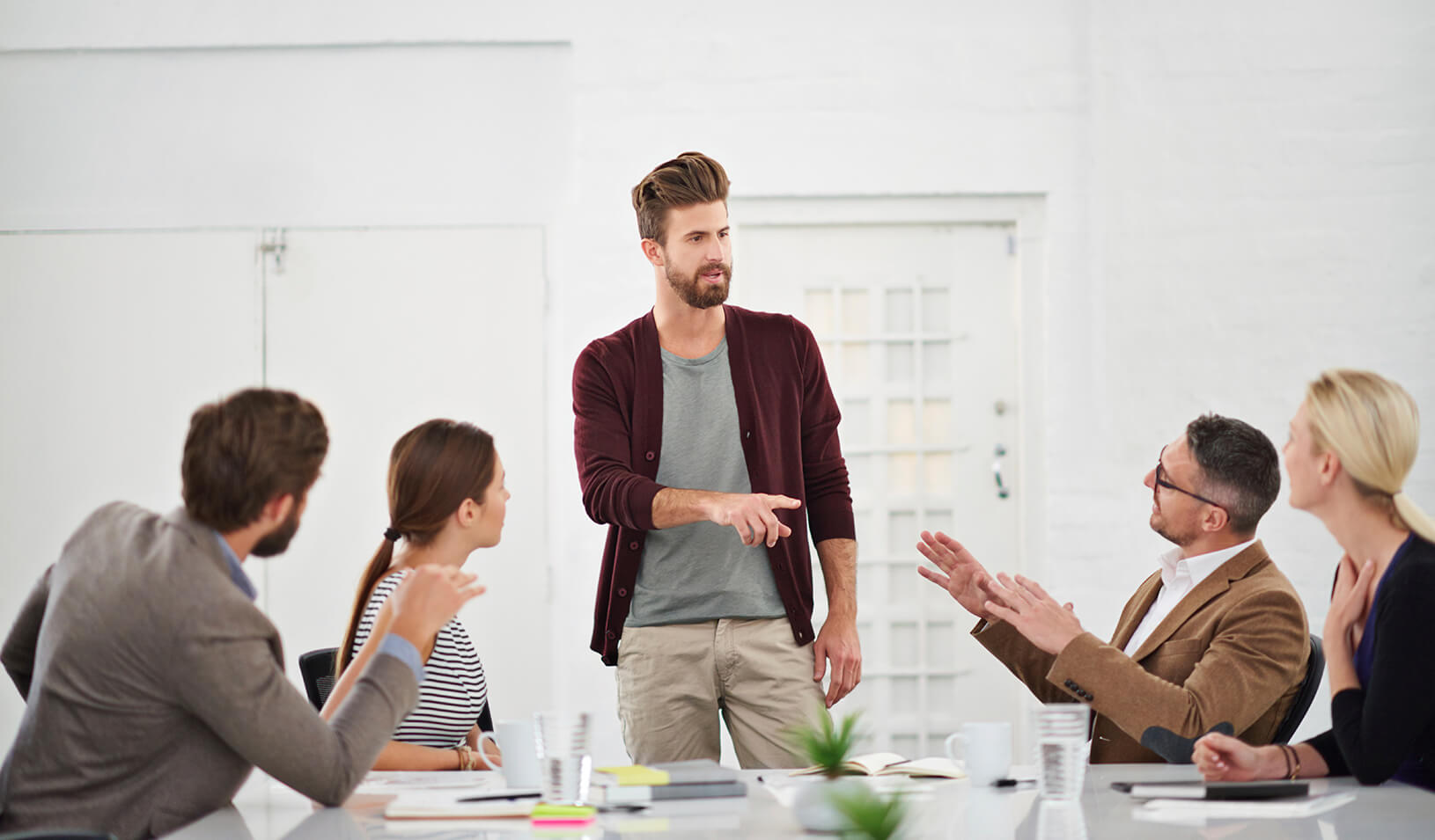 Twentysomething hipster standing while talking to a group of seated meeting participants | iStock/Yuri_Arcurs