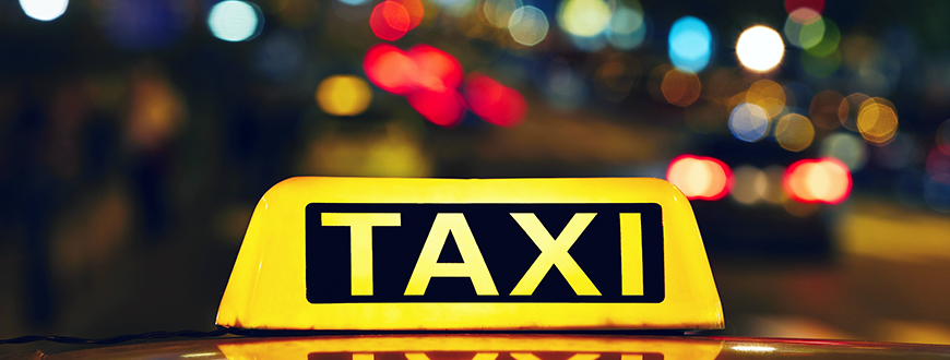 Yellow lit taxi sign on top of taxi, with building lights at night in background.