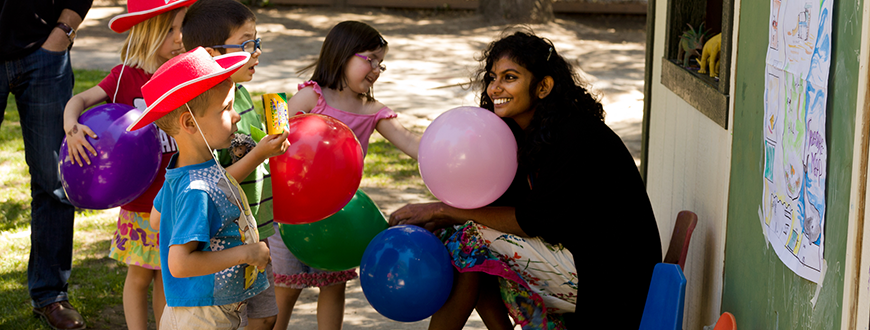 Two boys and two girls wearing hats and holding balloons, smiling and greeting Indian female.