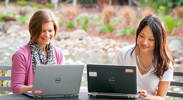 Two female colleagues smiling and looking at laptops, side by side on table otuside.