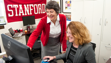 Two female colleagues smiling and looking at desktop computer screen with red Stanford banner in background.