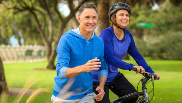 Couple in their 50s exercising in park; man is jogging, woman is biking.