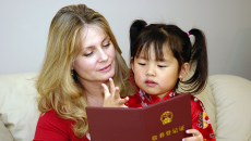 Caucasian mother reading a book with her Chinese daughter on the sofa.