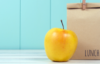 Brown lunch bag with yellow apple on table top against sky blue panelled wall.