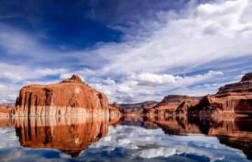 The blue sky and clouds reflect off the calm water in a reservoir surrounded by red rocky cliffs and outcrops. 