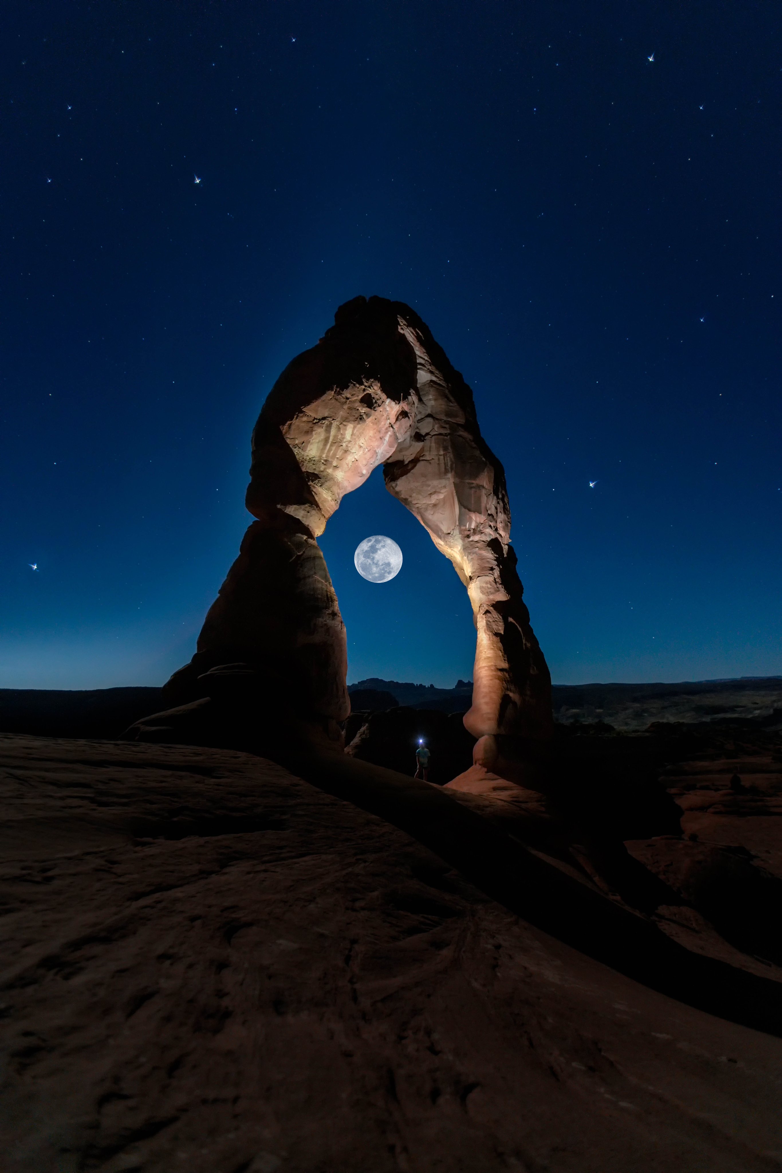 A large stone arch rises from layered rock as a full moon glows underneath.