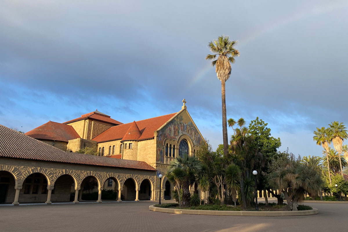 Empty Stanford quad with rainbow arching behind a palm tree.