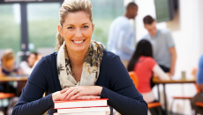 Mature female student smiling with hands on stack of books, in classroom with other students