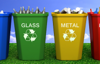 Four recycling bins on grass: blue for paper, green for glass, yellow for metal, red for plastic.