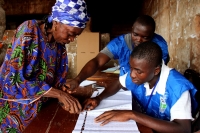 A woman casts her vote in the 2012 Sierra Leone elections by pointing to her chosen candidate while two men sit at a table displaying the elections ballot. 
