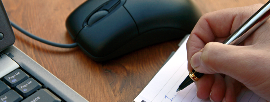 Closeup of person's hand taking notes next to laptop and computer mouse.