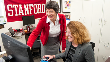 Two female colleagues looking at computer monitor. Stanford banner on wall behind them.