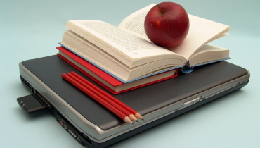 Open book stacked on top of laptop with pencils and red apple