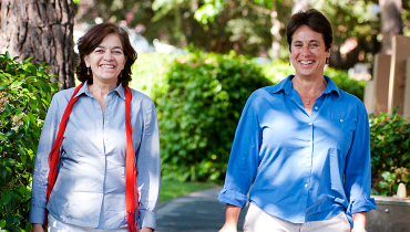 Two female staff members smiling and walking outside past trees and greenery.