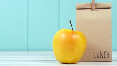 Brown lunch bag with yellow apple on table top against sky blue panelled wall.