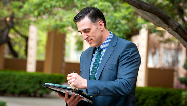 Male staff member in business suit writing on a padfolio, foliage outdoors in background.