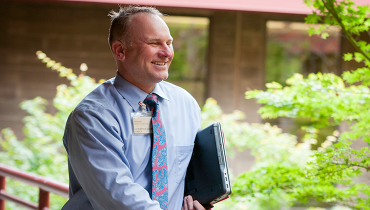 Photo of a Stanford employee smiling while holding a laptop with beautiful and lush greenery popping him out from the background. 