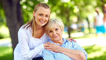 Young female holding hand of elderly female sitting in wheelchair outside in park.