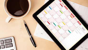 Calendar on tablet next to calculator, coffee cup and pen on desk