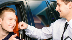 Male rental car rep giving car keys to male customer through driver seat window.
