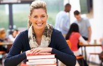 Mature female student smiling with hands on stack of books, in classroom with other students